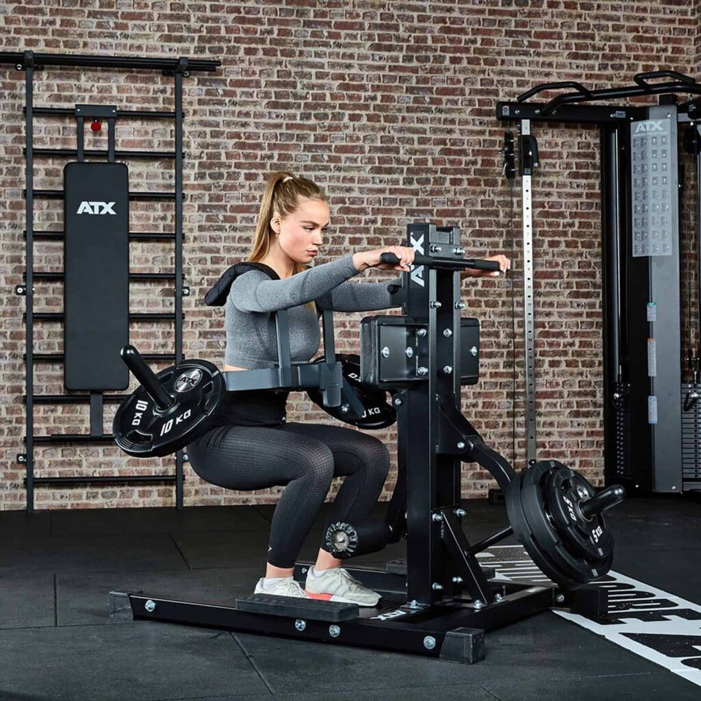 In a brick-walled gym, a person exercises on a seated calf raise machine with focus and determination, wearing workout gear. They are surrounded by various equipment, including the versatile ATX® Leg Master for comprehensive lower body strengthening.