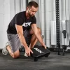 A man in a black ATX shirt and gray shorts kneels on a gym floor, placing an ATX® Seated Row Seat on the ground among various fitness equipment like a rack and weights.