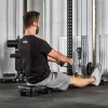 A person in a black ATX shirt and gray shorts is using the ATX® Seated Row Seat on a cable machine for rowing exercises. Theyre indoors, amidst gym equipment, facing away from the camera.