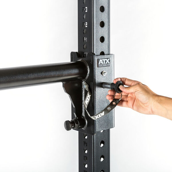 A hand adjusts a safety lock on the black metal ATX® Bulls Strongman Yoke, now discontinued. The rack, reminiscent of a strongman yoke, features multiple height-adjustment holes and a robust design against a white background.