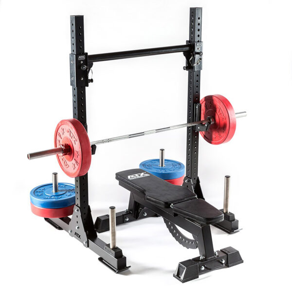 The ATX® Bulls Strongman Yoke DISCONTINUED features a robust setup with a weight bench and squat rack. A barbell, adorned with red and blue plates, mimics the strength of a strongman yoke. The bench beneath the barbell and extra plates stored on the rack base are set against a plain backdrop.