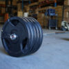 The Power Maxx 6ft Olympic Barbell, stacked with multiple 25 kg plates on both sides, sits on the gym floor. The backdrop features shelves and assorted equipment, highlighting a focused weightlifting or fitness area.