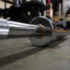 A close-up shows the 7ft Olympic Barbell Thrust Bearings 1500lbs resting on a gym floor. The shiny metal finish of the barbell end reflects the light, while blurred gym equipment and weights fill the background.