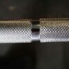 A close-up of the 7ft Olympic Barbell Thrust Bearings 1500lbs highlighting its textured knurling and contrasting shiny metal band against a dark background.