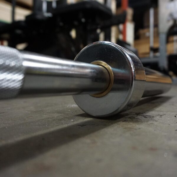 A close-up of the 7ft Olympic Barbell Thrust Bearings 1500lbs resting on a gym floor, focusing on the knurled grip and weight sleeve, with its shiny surface highlighted. Blurred in the background are gym equipment and storage shelving.