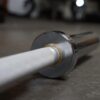 Close-up of the Aluminium 7.5kg Technique Olympic Bar resting on a gym floor. The textured grip of the metal bar contrasts with its gleaming, smooth ends. The focus is on the end part of the bar, with the background slightly blurred.