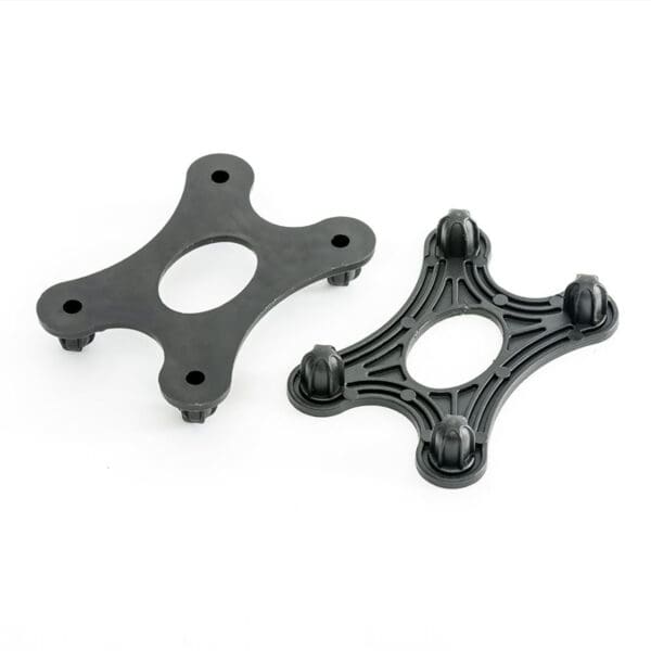 Two black, four-pronged GymFloor® RTS Turf components with oval centers are shown. One is solid with a smooth surface; the other has a grooved design and knobs at each tip. Both sit on a white background.