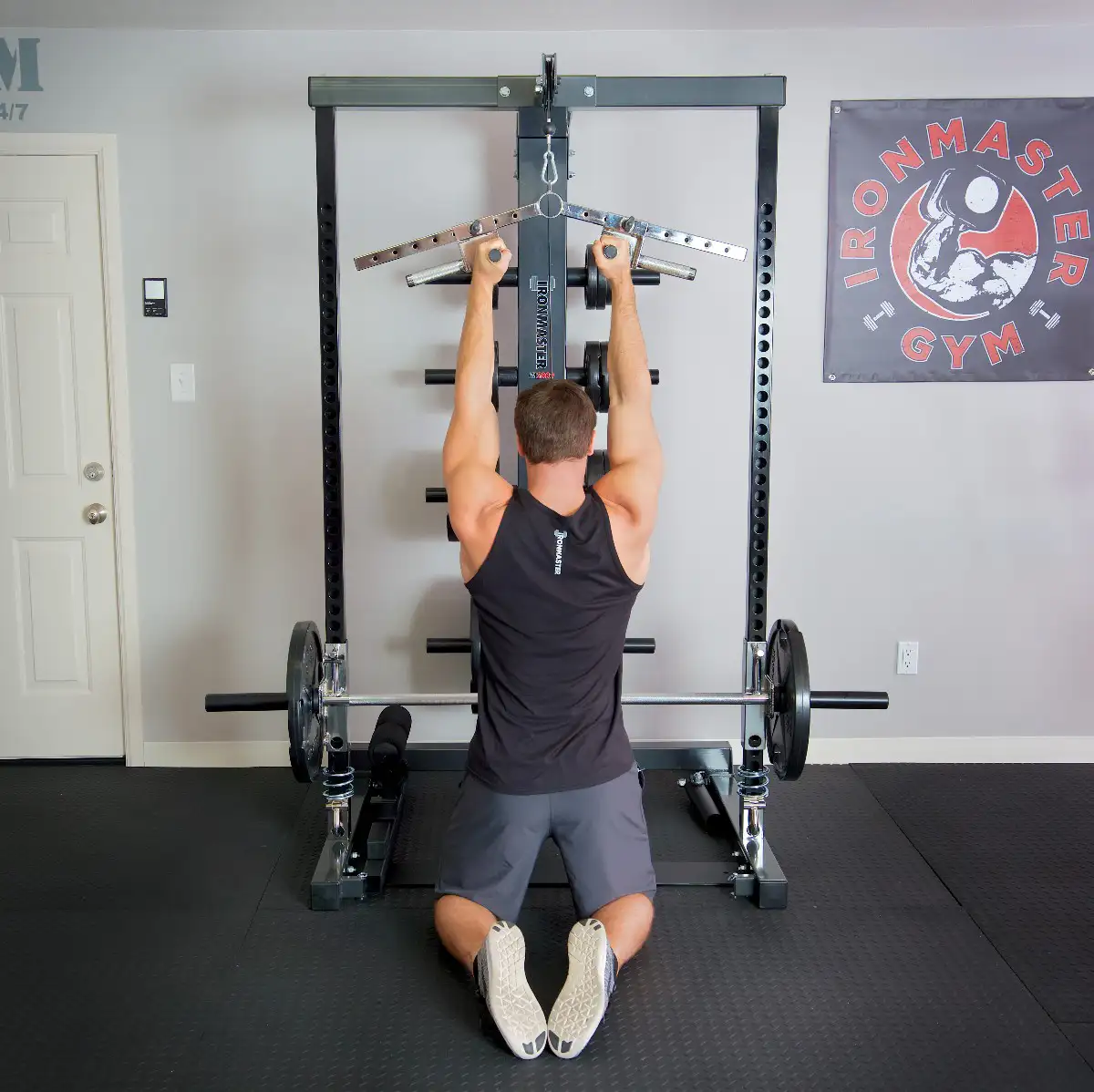 A man in a black tank top and gray shorts kneels on a mat, using the Ironmaster Ultimate Row Handle to perform a lat pulldown on a home gym machine. An Ironmaster Gym sign appears on the wall to the right.