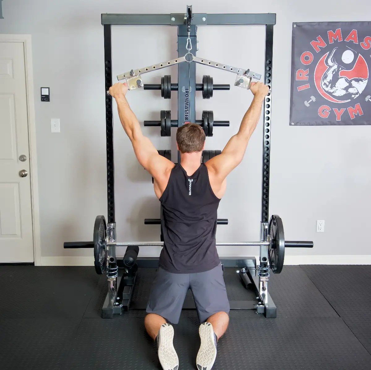 A man in a black tank top and gray shorts kneels while performing a lat pulldown with the Ironmaster Ultimate Row Handle using a cable machine in a gym, with weight plates and an IRONMASTER GYM banner on the wall.
