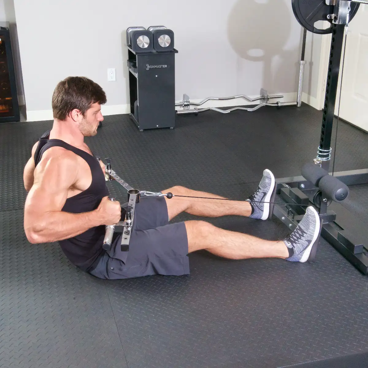 A man performs a seated row exercise in his home gym, pulling the Ironmaster Ultimate Row Handle toward his torso while wearing a black tank top, gray shorts, and athletic shoes.