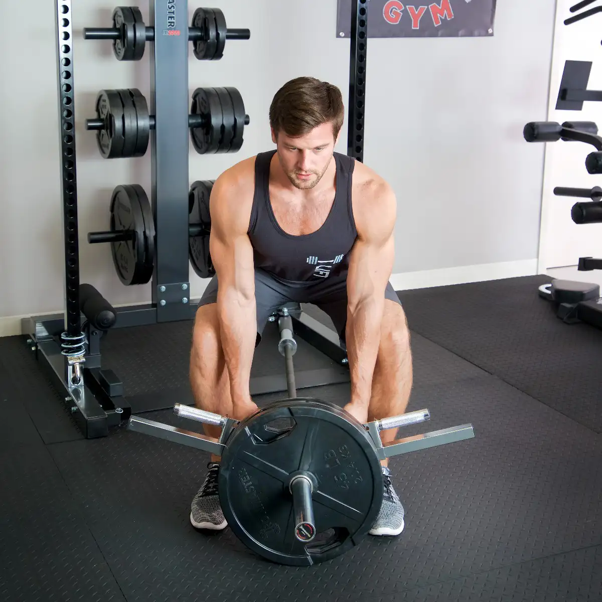 A man in athletic wear uses the Ironmaster Ultimate Row Handle to perform a T-bar row with a barbell in a gym, gripping the handles and lifting weights while standing on a rubber floor. Weight plates and gym equipment are visible in the background.