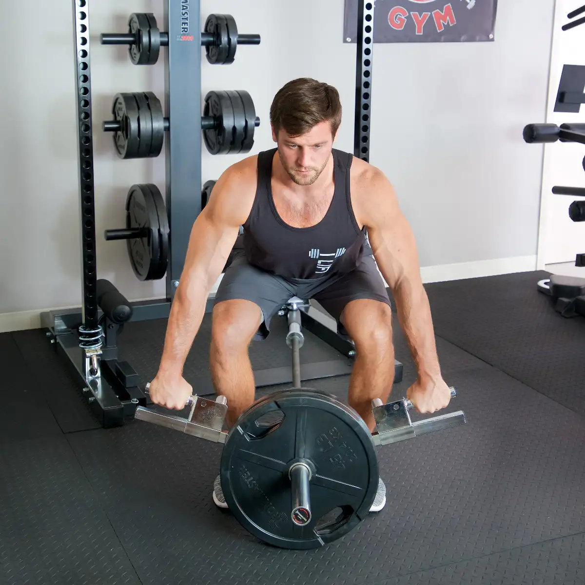 A man in athletic wear prepares to lift using the Ironmaster Ultimate Row Handle in a gym, surrounded by weight plates and workout equipment.