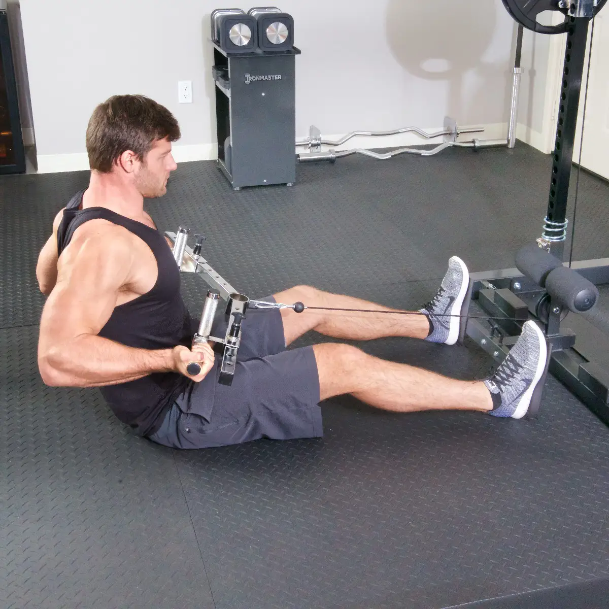 A man uses the Ironmaster Ultimate Row Handle in a gym, performing a seated cable row with straight back and bent knees, pulling the handle toward his torso.