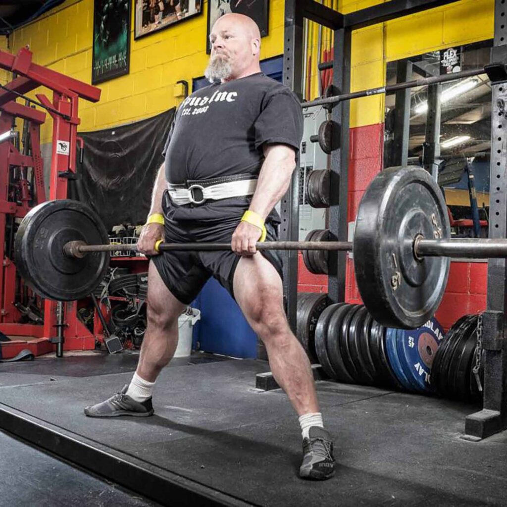 In a vibrant gym with colorful walls, someone is deadlifting in a black t-shirt, shorts, and weightlifting belt while wearing Spud Elbow Sleeves. Weight plates are neatly stacked behind them.