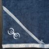A close-up of the Sting Microfibre Gym Towel in dark blue, featuring a diagonal light blue stripe and a white embroidered logo with two stylized curved lines. The bottom edge repeats the word STING along the border.
