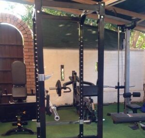 A home gym setup with a power rack, weight bench, and various gym equipment on a green floor. Its under a wooden roof with white walls and a brick archway entrance.