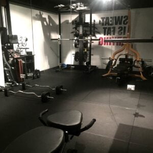 A dimly lit gym with weightlifting equipment, including benches, barbells, and a squat rack. A motivational quote on the wall reads, Sweat is fat crying. The floor is covered with black rubber mats.