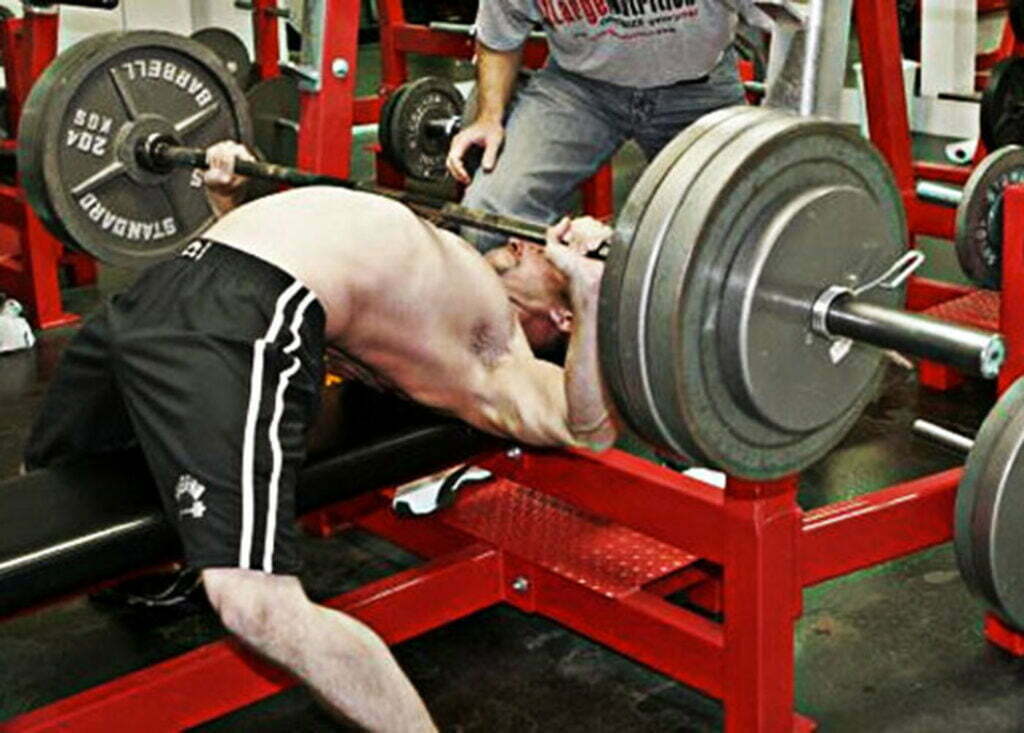 A person performing a bench press in a gym, with a spotter standing nearby. The barbell is loaded with several weight plates. The lifter is wearing black shorts, and the equipment is red.