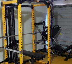 A yellow power rack equipped with a bench, barbell, and pulley system in a gym setting. Theres also a leg press machine visible in the background. The gym has a concrete wall and a partially open garage door.