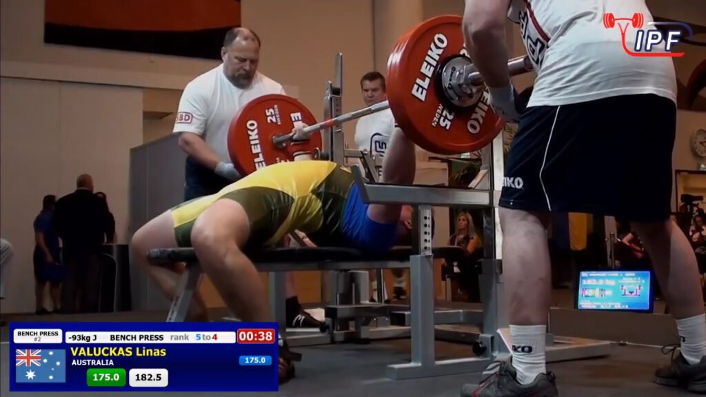 A weightlifter in a yellow and blue uniform performs a bench press during a competition. Two spotters stand by for safety. Digital displays show the weight as 175.0 kg and the athletes details. A crowd watches in the background.