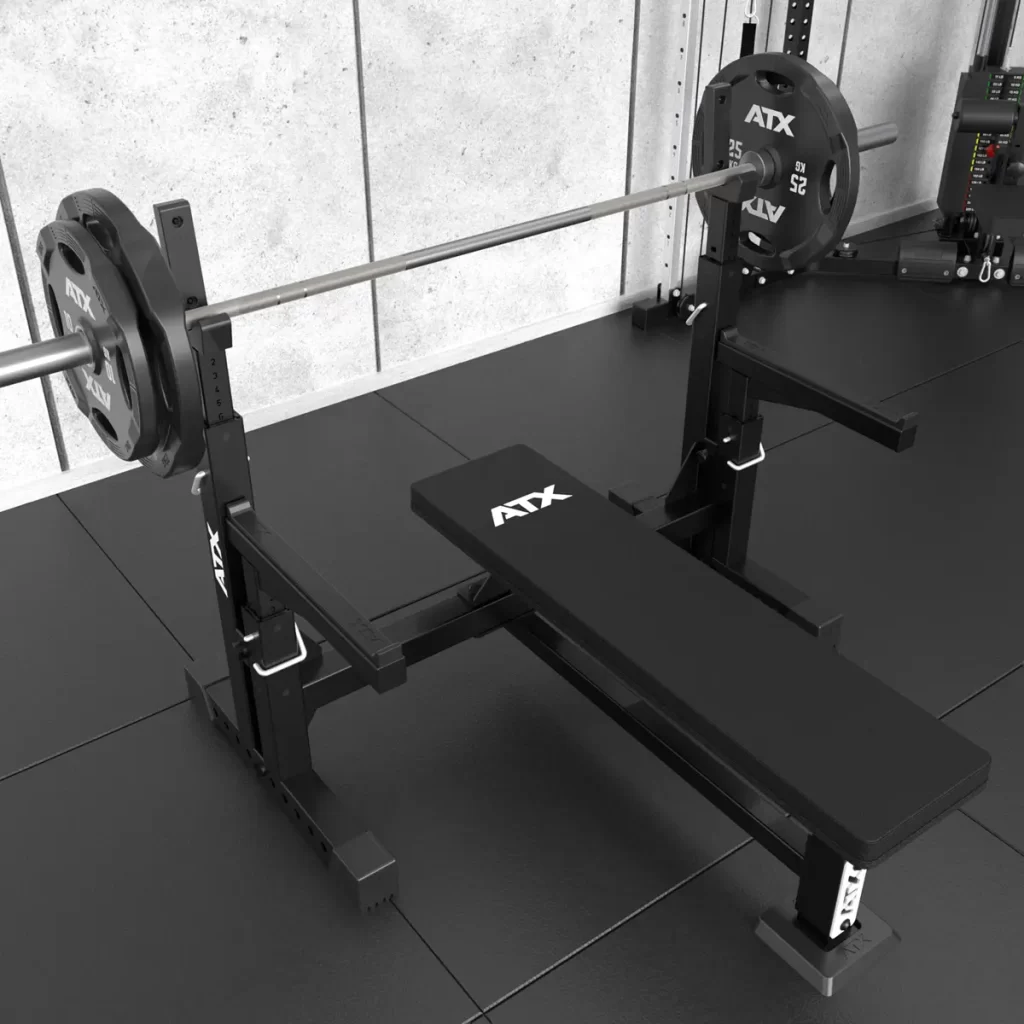 A black ATX® Olympic Bench Press 700 with a barbell and two large weight plates on each side is situated against a gray concrete wall in a fitness facility.