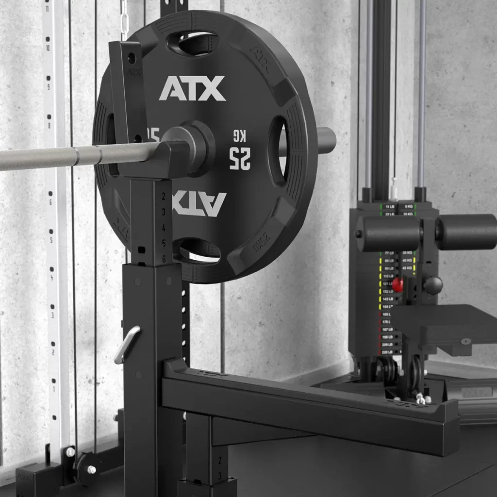 A gym setup with an ATX® Olympic Bench Press 700 loaded with a 25 kg ATX weight plate on a squat rack, surrounded by adjustable safety bars and a cable machine, against a concrete wall backdrop.