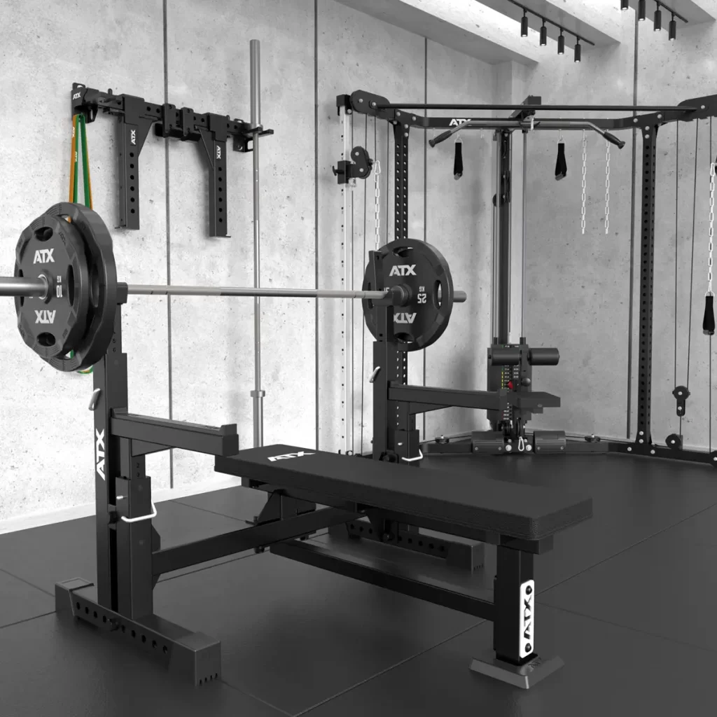 A modern gym equipped with the ATX® Olympic Bench Press 700 setup, featuring a barbell and weights. In the background, theres a multifunctional cable machine, wall-mounted resistance bands, and black rubber flooring.