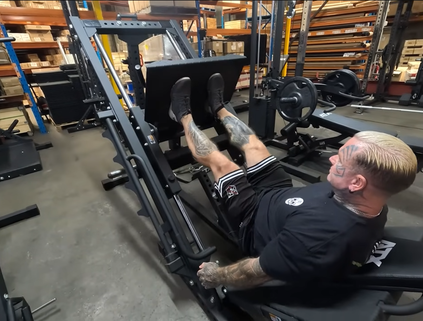 A heavily tattooed person with blond hair is using a leg press machine in a gym surrounded by various weights and equipment. They are lying back, pushing a weighted platform with both feet.