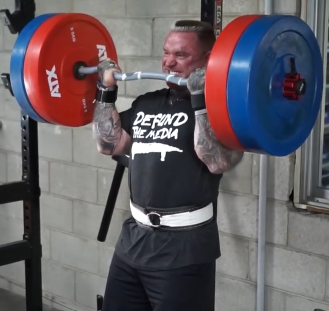 A muscular person with tattoos lifts a heavily loaded barbell in a gym, wearing a DEFEND THE MEDIA shirt and a weightlifting belt, straining with effort.