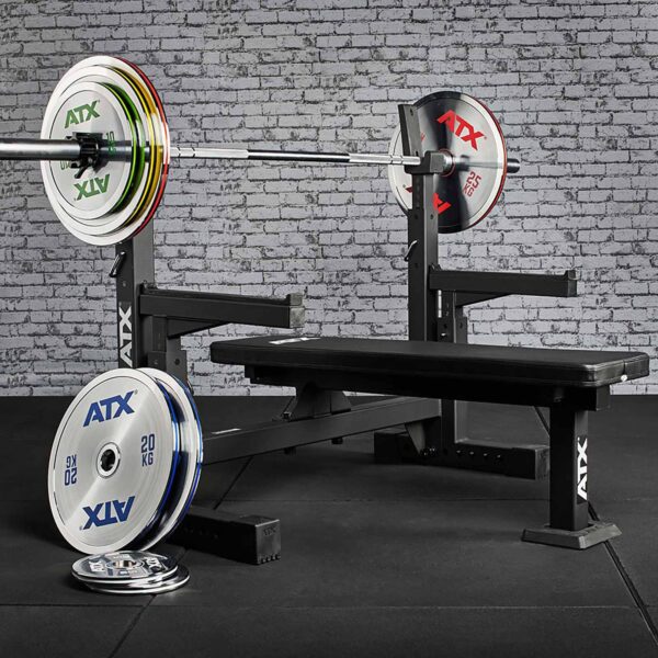 A gym setup showcases a bench press with a barbell loaded with ATX® 25kg Chrome Powerlifting Single Plates. Plates are neatly stacked on stands and the floor. The equipment rests on a black mat against a brick wall, crafting a professional workout space.
