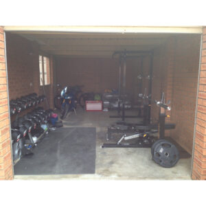 The image shows a garage partially open, revealing a home gym setup with a squat rack, weight plates, and dumbbells on the left. On the right, a motorcycle is parked near a red toolbox against the brick walls.
