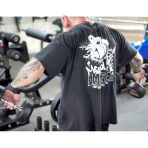 A person with tattoos is lifting dumbbells in a gym. They are wearing a black t-shirt with a graphic design on the back. Gym equipment is visible in the background.