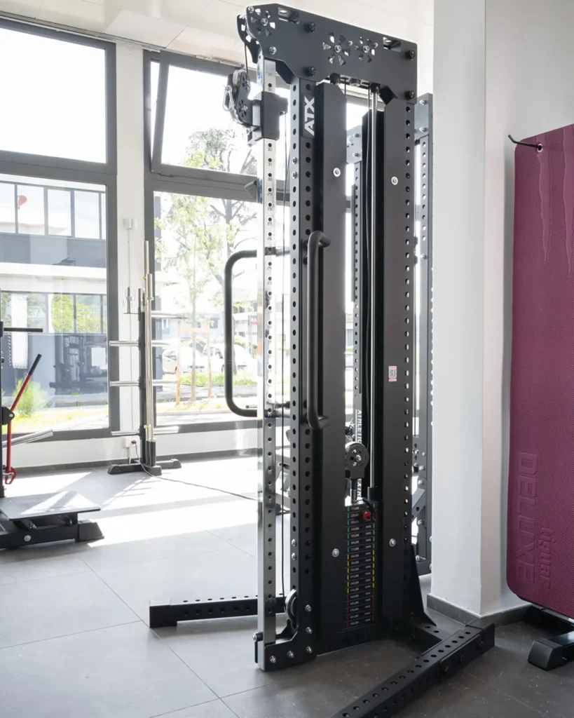 In a bright gym with large windows, a modern cable weight machine stands elegantly. The sleek, black equipment, part of the main cable station, features adjustable weights and metal handles. Sunlight streams in, casting shadows on the tiled floor.