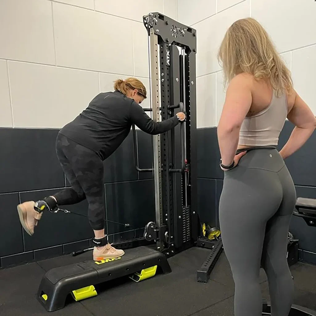 A person exercises on a cable weight machine, balancing on one leg with adaptive equipment on the other. A trainer in a tank top and leggings observes. They are in a gym with a black floor and white walls.