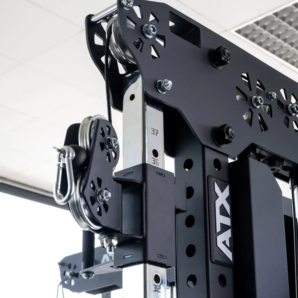 Close-up of a black and silver cable weight machine with visible cables and metal components. The equipment is marked with numbers and a logo. Bright indoor lighting highlights the precision and design of the machinery.