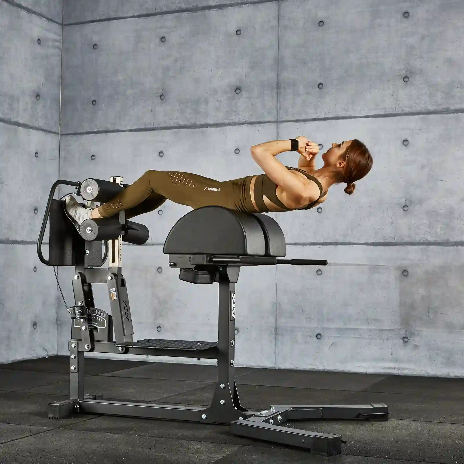 A person in workout attire is performing an exercise on a Roman chair, resembling the style of a glute and ham raise machine, against a gray concrete wall in the gym. They are reclined with legs secured and arms crossed over their chest.