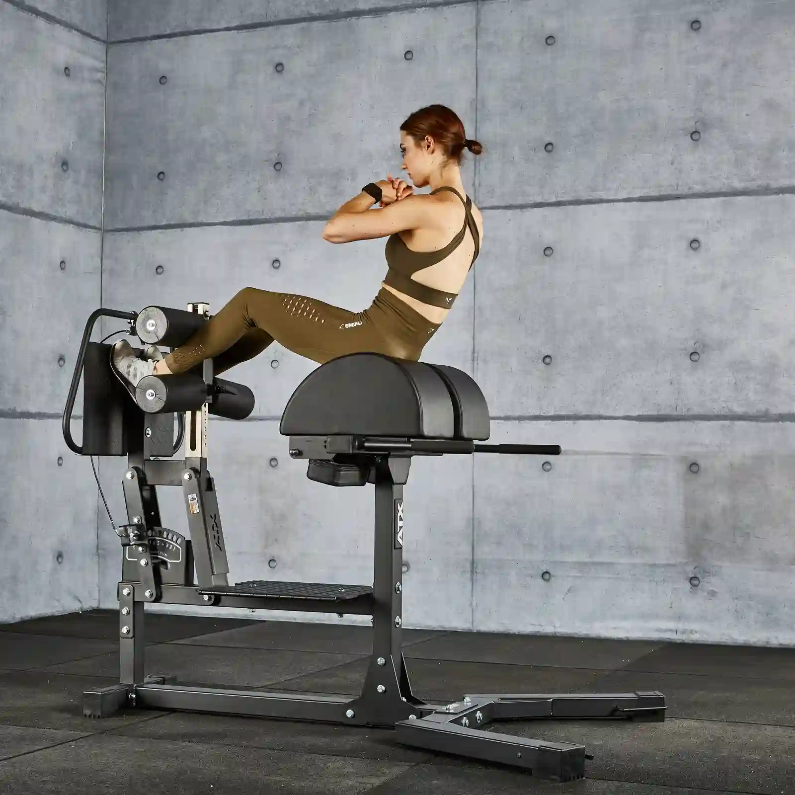 A person in athletic clothing performs a back extension exercise on a GHD gym machine in a concrete-walled gym.