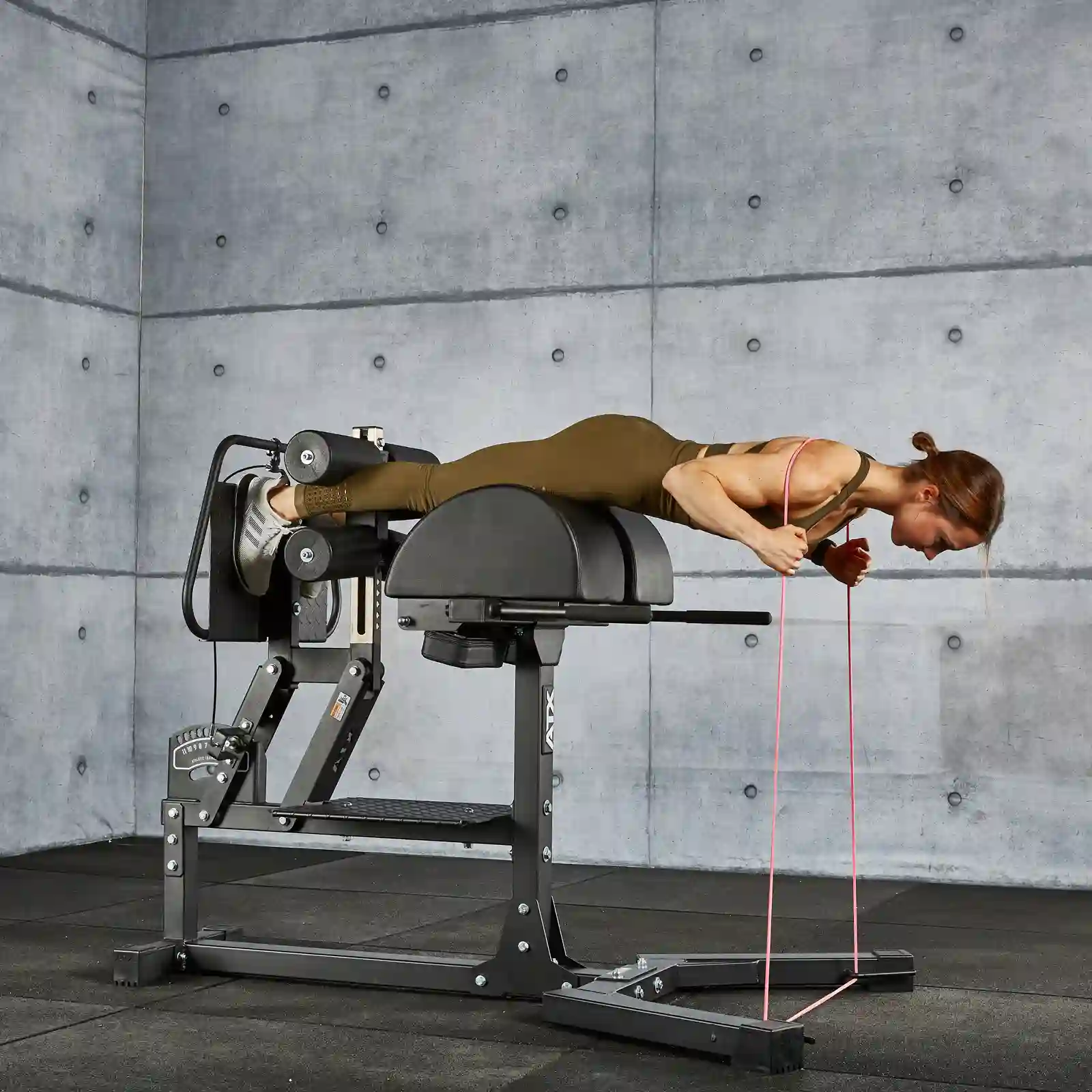 A person in athletic clothing performs a back extension exercise on a hyperextension bench in a gym. The walls are made of gray concrete blocks. A resistance band is attached to the bench and the floor is covered with black mats, right next to the GHD gym machine.
