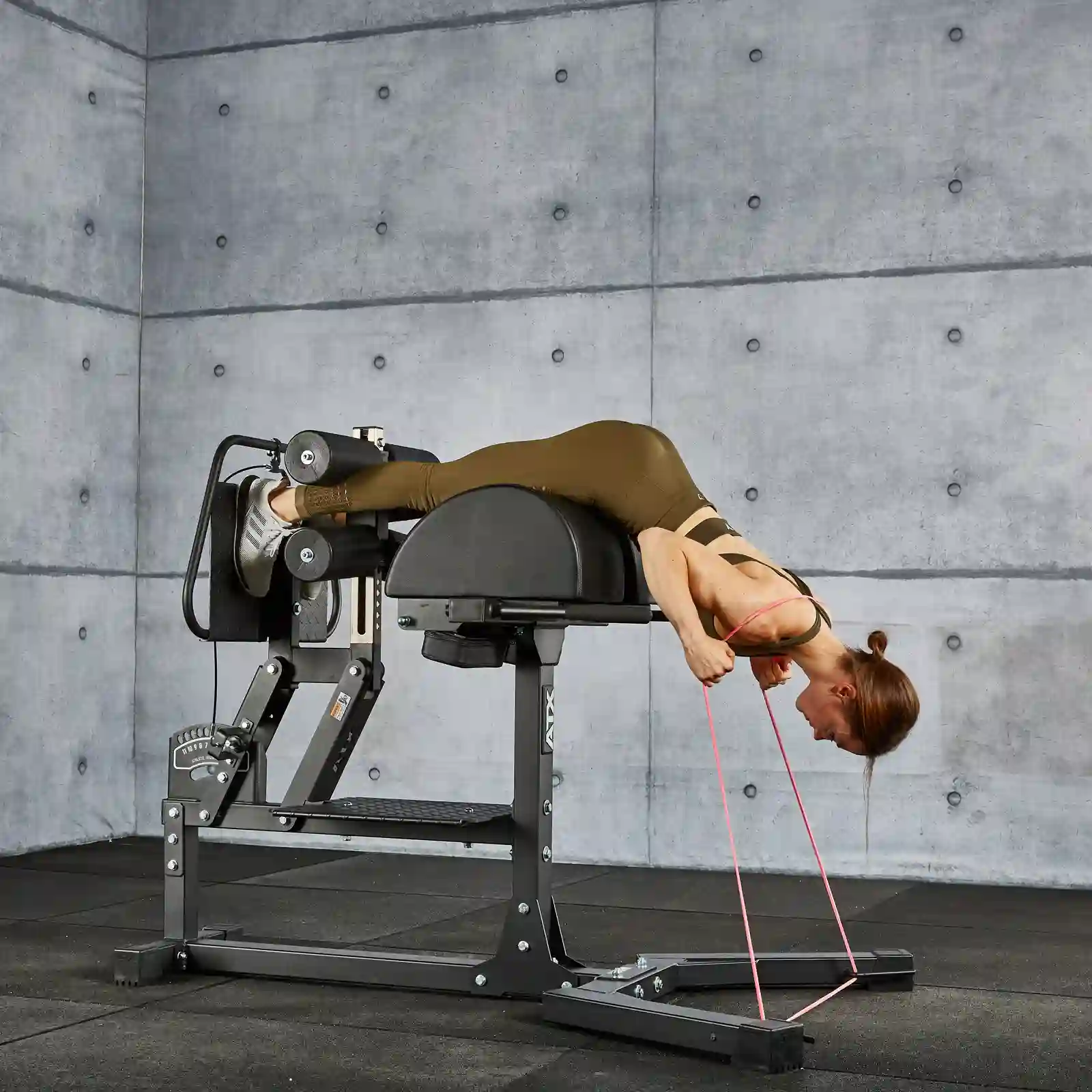 A person in brown workout attire is performing a back extension exercise on the glute and ham raise machine, utilizing a resistance band for added difficulty. The gym has concrete walls and black flooring.