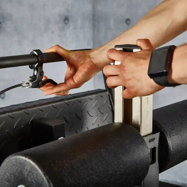 A person adjusts the handles of a cable machine in a gym, preparing for their glute extension exercises. They are wearing a smartwatch on their wrist. The machine features black padded support and a metal weight stack in the background, ideal for targeting lower body muscles.