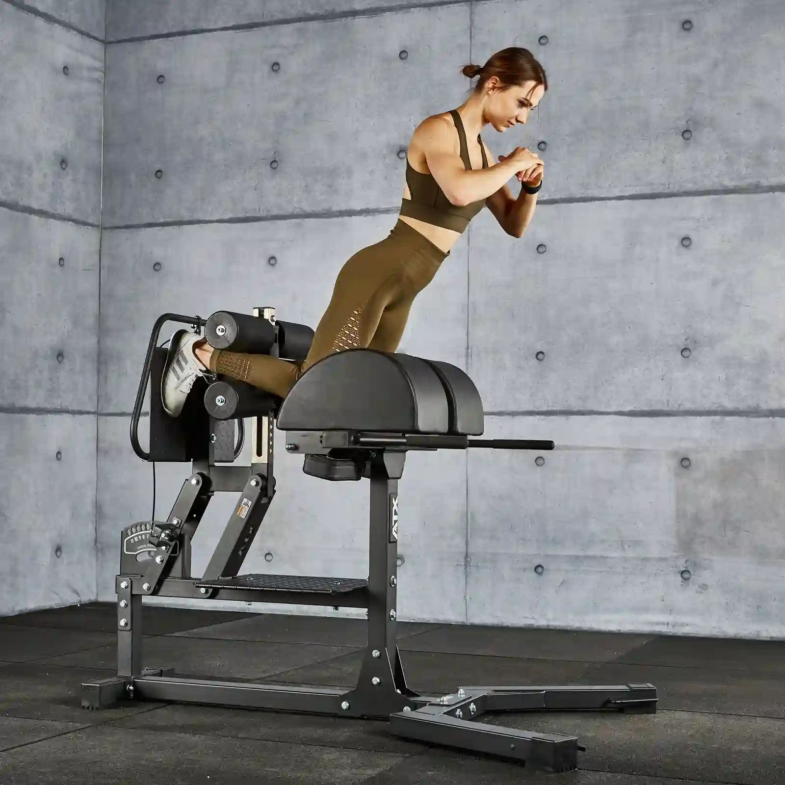 A woman exercises on a glute and ham raise machine in the gym. She wears a brown sports outfit and checks her smartwatch during her workout. The background features a gray concrete wall, adding an industrial chic vibe to the scene.