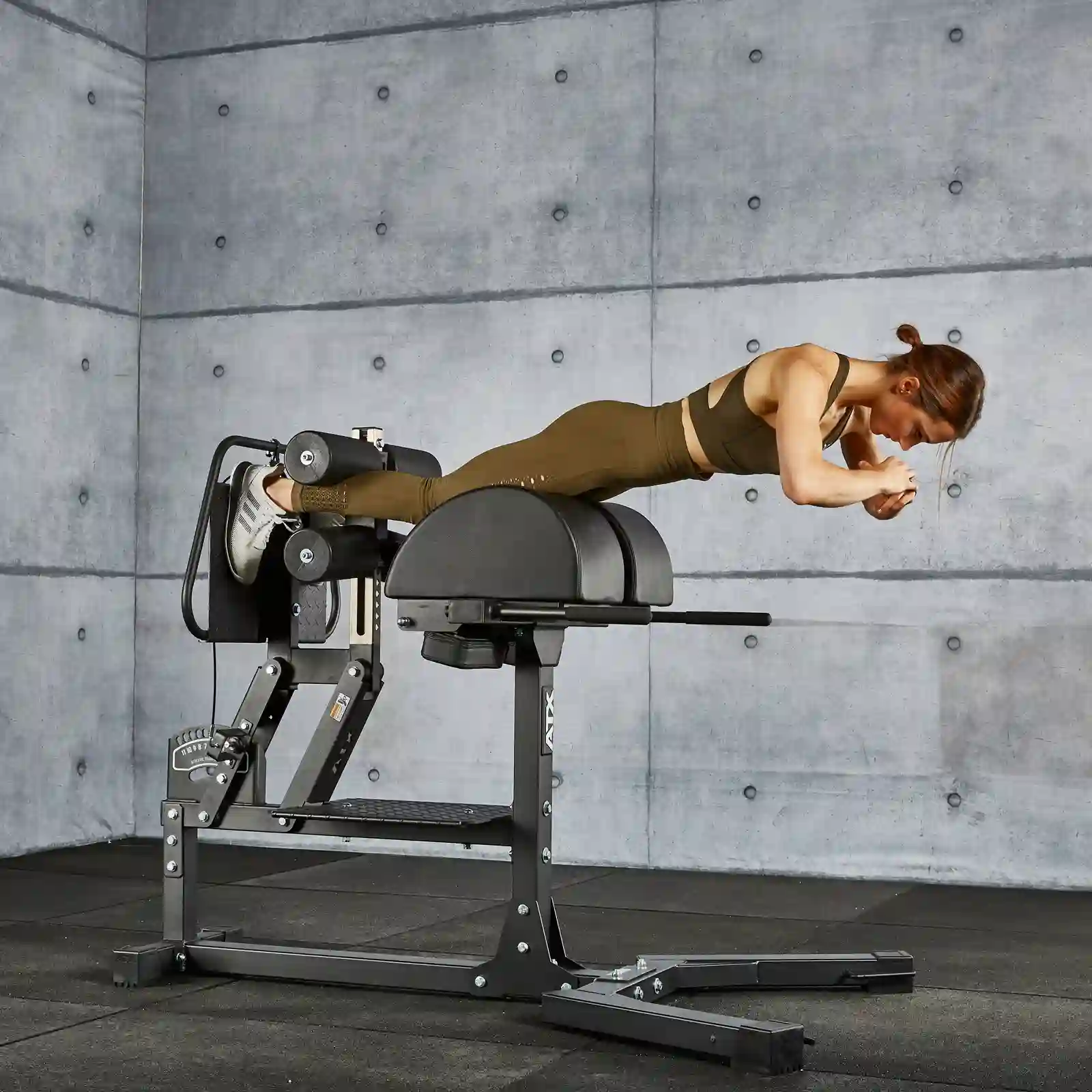 A person in workout attire uses a GHD gym machine in a gym with gray concrete walls. They are positioned horizontally, engaging in the exercise.