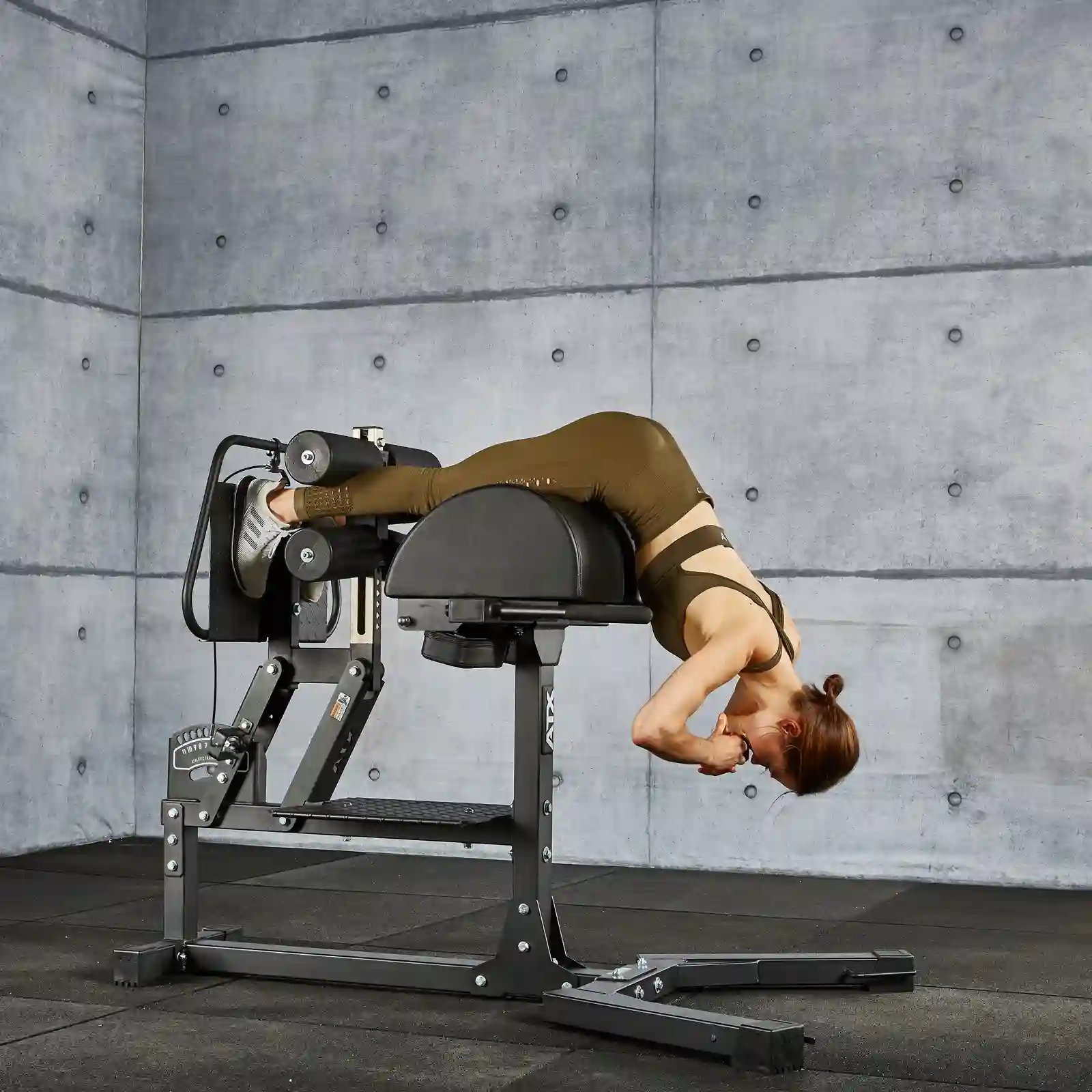 A person in athletic clothing performs a back extension exercise on a GHD machine in a gym with gray walls and black flooring.