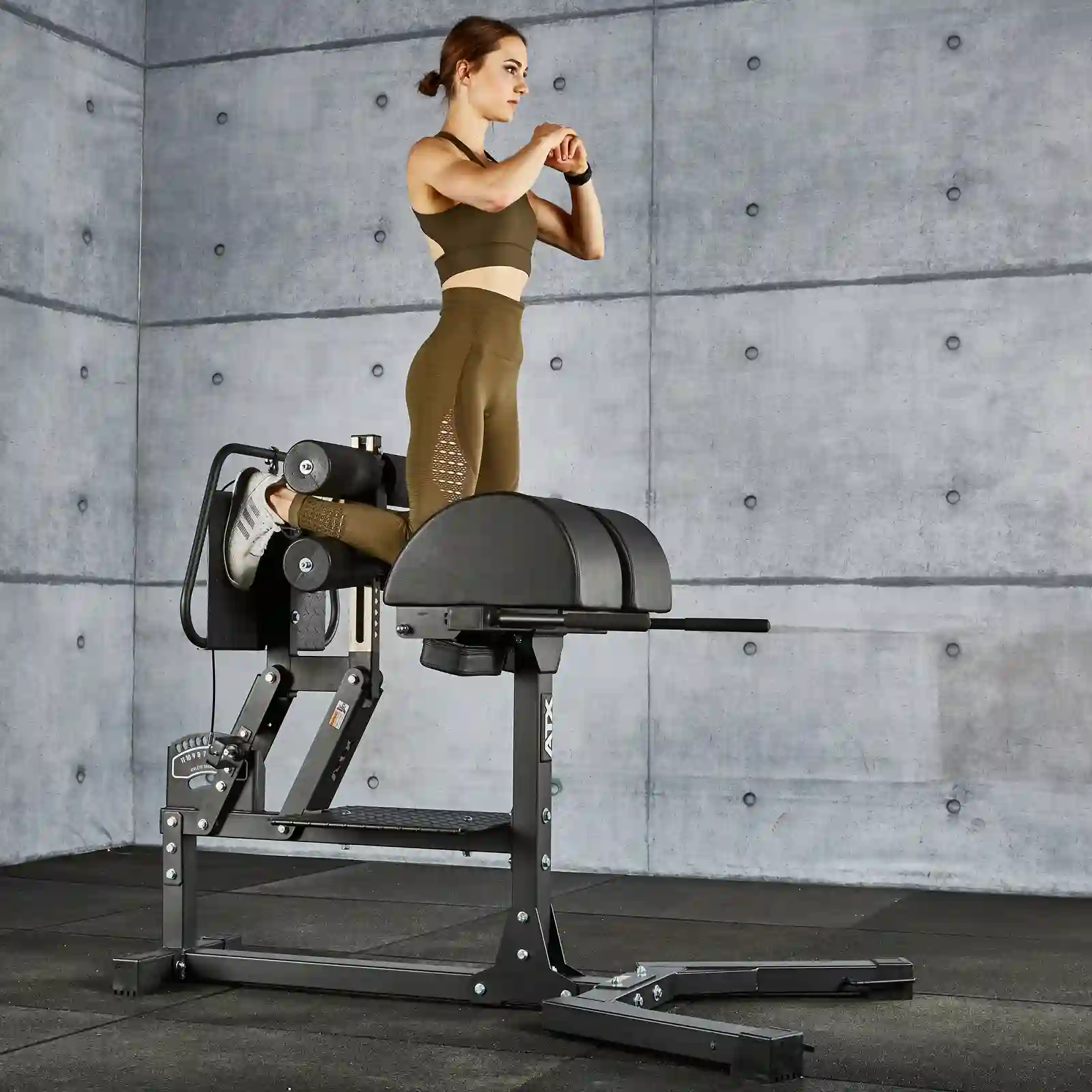 A person in workout gear is using a Roman chair hyperextension machine, also known as a GHD machine, in a gym. They are positioned at an angle, with arms folded in front of their chest, performing a back extension exercise against the concrete wall backdrop.