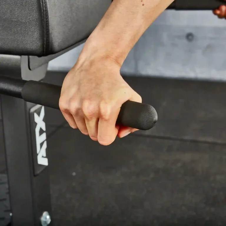 A person's hand grips the black foam handlebar of a glute machine positioned on a dark gym floor. The firm grip indicates that the user is engaged in an intense exercise routine, focusing on strengthening their lower body.