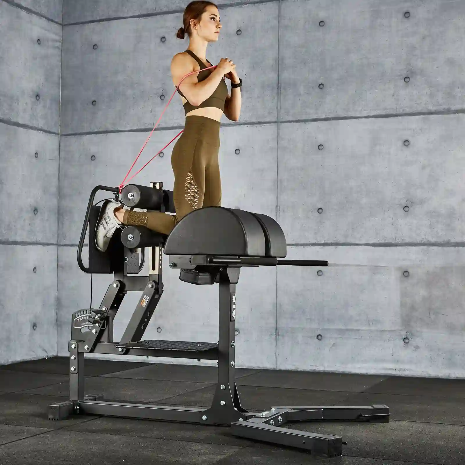 A woman in athletic wear utilizes a glute and ham raise machine, performing a precise back extension with resistance bands attached. The modern gym setting, complete with concrete walls, complements her focus on the glute extension workout.