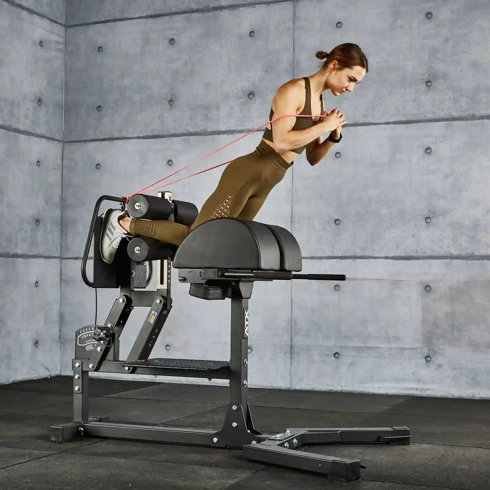 A person is using a hyperextension bench in a modern gym, effortlessly performing exercises with a resistance band and targeting their muscles like on a glute and ham raise machine. Clad in a brown workout outfit, they focus amid the sleek concrete walls and black flooring.