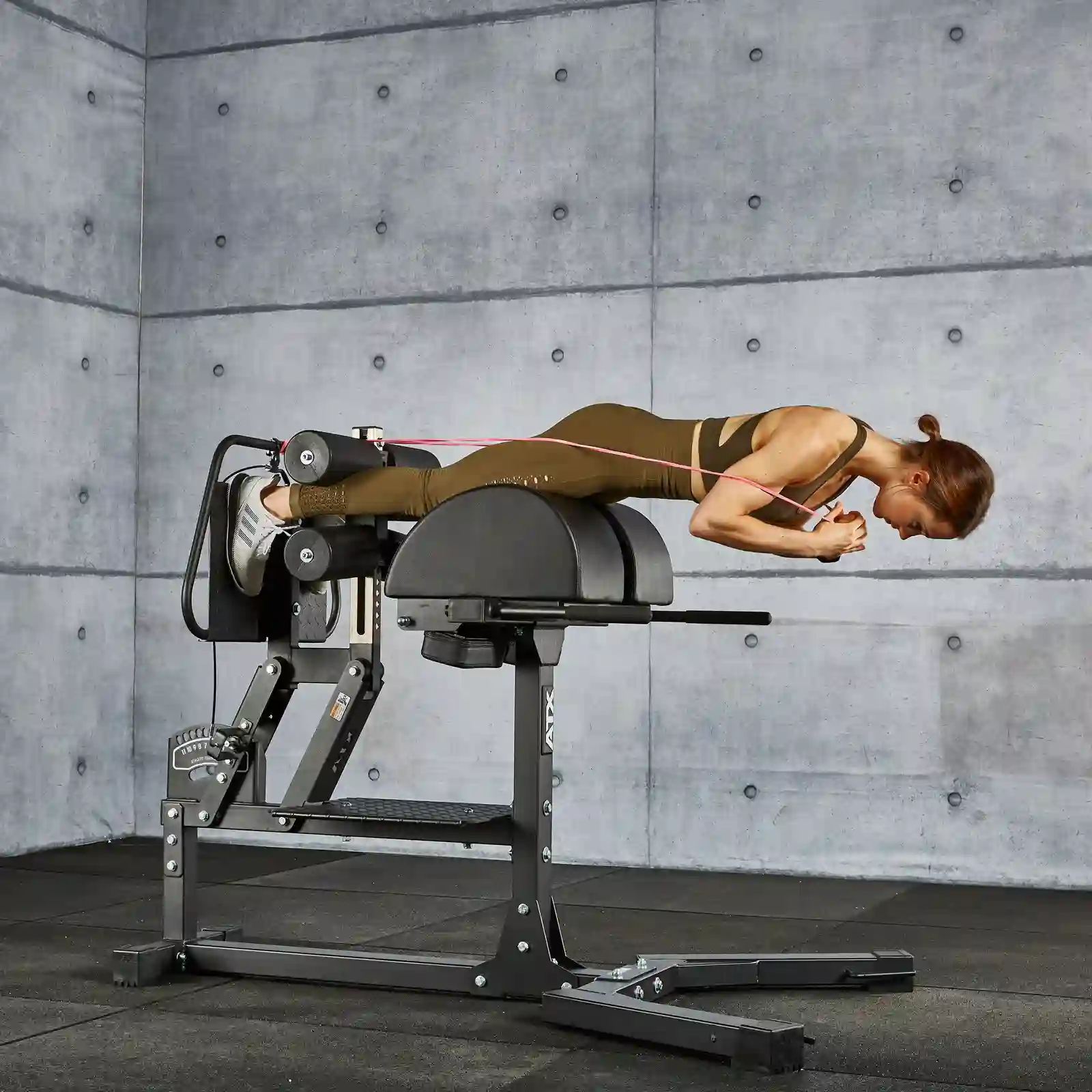 A person exercises on a hyperextension bench, performing back extensions beside a ghd gym machine. The room has a gray concrete wall and black floor tiles. They’re wearing a brown workout outfit and white shoes.