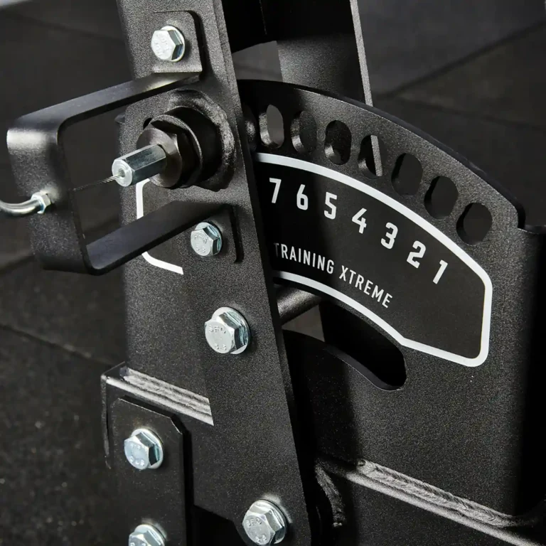 Close-up of a black metal exercise equipment adjustment mechanism, likely from a glute and ham raise machine. It features a numbered dial from 1 to 7 and the text TRAINING XTREME. Bolts and a pin are visible, set against a dark background.