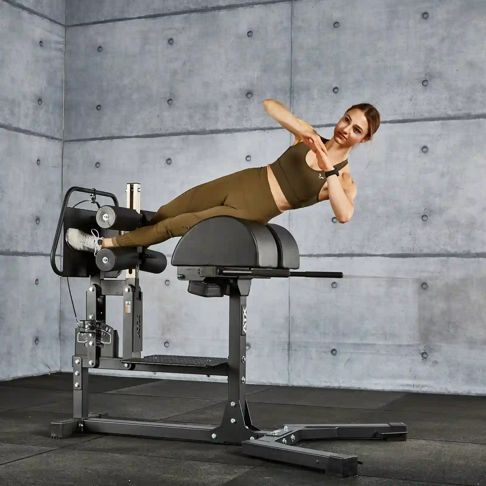 A person in workout attire performs a side plank on a Roman chair in a modern gym with a textured gray wall. Nearby, the glute and ham raise machine stands ready for action. They are balancing with one arm across their chest and the other on their waist.
